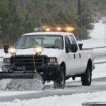 A pickup with a yellow light bar and a plow attachment clears a road of snow, 背景是松树.